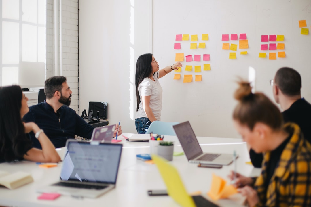 Five individuals share a conference room, viewing laptops and sticky notes as they plan a project.