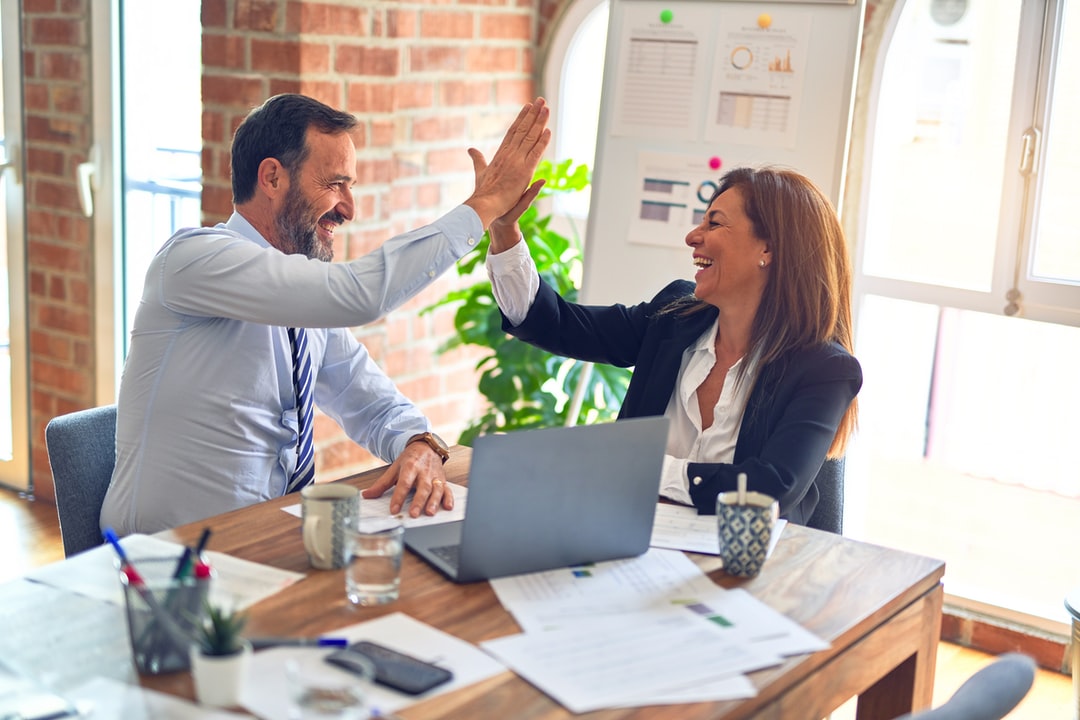Alt text: A man and woman dressed in professional attire high-five and smile over a desk cluttered with a computer and papers.